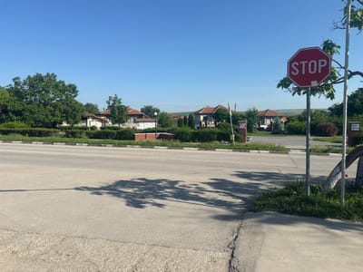 The image depicts a street scene in Tsenovo, Bulgaria. A "STOP" sign is prominently displayed on the right side of the road. A residential area with houses having red-tiled roofs is visible in the background. The sky is clear and blue. A shadow of a tree falls across the road. There are trees and greenery along the street. A sign is barely visible to the right of the stop sign on a tree.