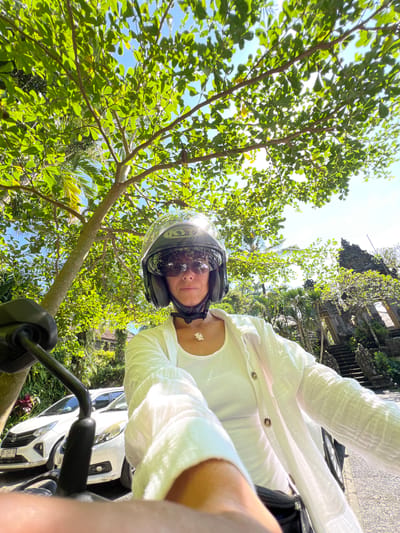 A first-person, upward-angled, eye-level outdoor shot depicts a person wearing a motorcycle helmet and sunglasses on a motorbike under trees. The person wears a white button-down shirt. A white flower pendant necklace is visible. The handlebars of the bike are in the foreground. A white car is partially visible on the right side. The trees are lush with green leaves, and the sky is blue with sunlight filtering through the leaves. In the background is traditional Balinese stonework, indicating the location is Kedewatan, Indonesia.