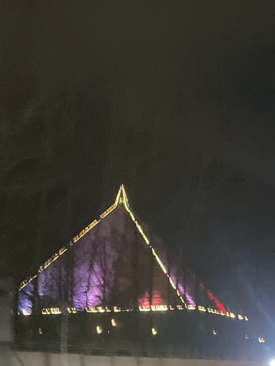 A night shot shows a large triangular building with illuminated edges in Verin Ptghni, Armenia. The building's reflection is visible, casting purple hues. There is a dark area in the left portion of the image.