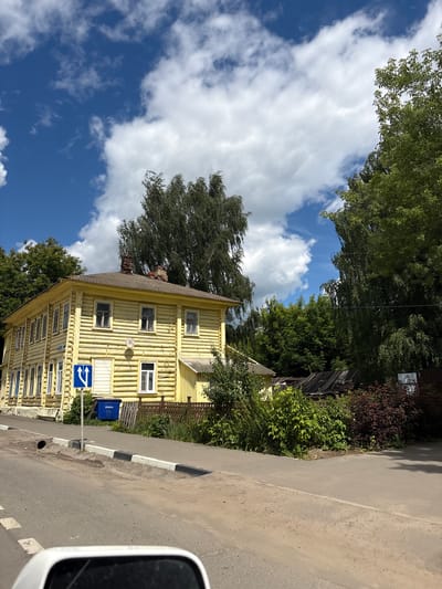 This is a vertical, eye-level, outdoor shot of a yellow, wooden, two-story building situated next to an asphalt road. The building is constructed from logs and has numerous windows with white frames. A blue sign with white arrows indicating a mandatory direction of travel (left and right) is visible in front of the building. A blue trash receptacle is also present near the building. The scene is framed by lush green trees and a bright blue sky with scattered white clouds. The lighting suggests it is daytime, likely sunny. The image has a rustic feel, possibly indicating a rural or historical setting. A black and white striped curb runs along the edge of the road. The scene appears to be in Pereslavl-Zalessky, Russia. No people are visible in the image.