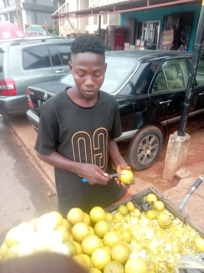 A young man stands behind a cart of citrus fruits in Umuokpu, Nigeria. He peels a yellow citrus fruit with a small knife. The cart contains numerous yellow citrus fruits and some white ones, along with shredded pieces of yellow peel. A black car and a silver SUV are parked behind him. The street appears wet. A building is partially visible in the background.