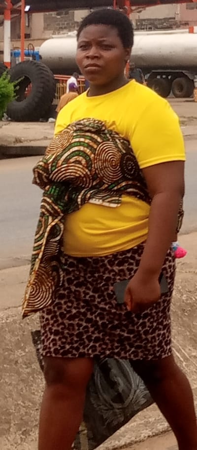 An individual with dark skin and short, dark hair stands outdoors in Awkuzu, Nigeria. This person is wearing a plain yellow t-shirt and a brown leopard-print skirt. A patterned fabric, featuring brown, green, and white circular designs, is draped over the individual's left shoulder and torso. A dark-colored smartphone is held in the right hand, positioned at the side. A small pink and white object is visible near the right hip, possibly tucked into the skirt or fabric. The individual faces forward with a neutral expression. In the foreground, a gray and black object or bag is partially visible on the ground near the individual's feet. The immediate background shows a road surface and a light-colored curb. Further back, a large black industrial tire is prominent on the left. To the right of the tire, a large white cylindrical tank, consistent with a tanker trailer, is visible. Multiple blurry figures are discernible in the mid-background. Structures and buildings are present in the far background.