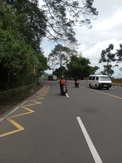 This image depicts a multi-lane asphalt road curving gently to the right, under a partially cloudy sky, located in the city of Boommlog, Bhutan. In the foreground, a dark-colored scooter is ridden by an individual wearing a red jacket and a helmet, moving away from the camera. Immediately behind this scooter, a red motorcycle is operated by another helmeted individual in dark attire, also moving away.
The road features a solid white line extending diagonally across the lower right foreground, which serves as a lane divider. On the left shoulder, yellow zigzag markings are painted on the asphalt, bordered by a solid white line and an adjacent concrete drainage ditch with dense green vegetation on a raised bank. In the opposing lane, a white van, appearing to be a Mitsubishi L300 model, is driving towards the camera. Further down the road in the opposing lane, a yellow truck is visible. A black and yellow striped barrier runs along the right side of the road, separating it from a vegetated slope. The background consists of a mix of broadleaf and conifer trees, with a utility pole or traffic signal structure visible in the distance.