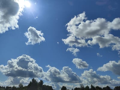 The image depicts a sky filled with scattered white and gray clouds against a clear blue sky. The sun is visible in the upper left quadrant of the image, casting a bright glare and lens flares. Below the sky, a line of dark green trees forms the horizon, suggesting an outdoor environment. The scene appears to be a landscape view of the sky over Stapriņi, Latvia. There are no people or specific objects of interest other than the natural elements of the sky and trees.