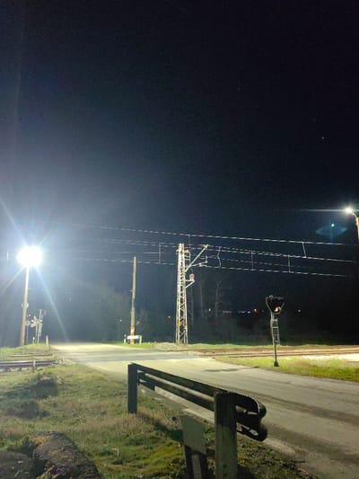 The image depicts a nighttime scene at a railway crossing in Nadarevo, Bulgaria. The environment is illuminated by artificial light sources, creating significant lens flare from two prominent overhead lights: one positioned on a pole to the far left, and another further right at the edge of the frame.
In the immediate foreground, a dark, weathered metal barrier with two horizontal rails extends from the bottom right towards the center, separating a grassy, uneven embankment from an asphalt road. The ground consists of sparse grass, exposed soil, and some debris.
The asphalt road traverses the midground, crossing two parallel railway tracks laid on gravel ballast. To the left of the crossing, a white crossbuck railway crossing sign is visible. A tall, thin wooden pole stands vertically near the road's edge on the left, behind which is a dense, dark treeline.
Further into the scene, centrally positioned between the road and the tracks, stands a metal lattice mast supporting the railway's overhead electrification (catenary) system, which includes multiple wires stretched across the width of the tracks. To the right of the tracks, a tall, slender railway signal post with a dark housing and a visible orange element (possibly a light or reflector) is present.
The background consists of a dark, indistinct treeline beneath a dark, clear night sky. No individuals, animals, or vehicles are present in the frame.
