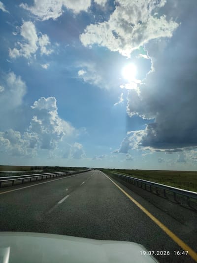 The image depicts a view from a vehicle traveling on a highway. The road is asphalt with white lane markings and a solid yellow line on the right side. Guardrails are visible on both sides of the road. The landscape on either side of the highway appears to be flat, grassy plains with sparse vegetation and some trees in the distance. The sky is a bright blue with a mix of white cumulus clouds and a large, dark storm cloud formation on the right. The sun is partially obscured by clouds, casting bright rays of light. In the distance, a few vehicles can be seen on the highway. A date and time stamp, "19.07.2026 16:47", is visible in the lower right corner of the image. The location is identified as Бәйет ауылдық округі, Kazakhstan. The scene captures a moment of travel on a highway in a rural, steppe-like environment under a dynamic sky.