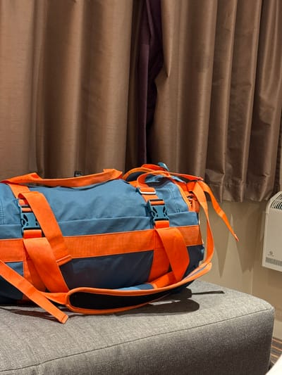 The scene depicts an interior setting, identified as being in the city of New Forest, United Kingdom. A large, approximately 60-70 cm long, cylindrical duffel bag is centrally positioned, resting on a grey, textured, upholstered surface. The bag is constructed from a teal-blue fabric and features prominent, bright orange webbing straps that run both longitudinally and transversely around its circumference. Multiple orange straps form carrying handles, and others are secured with teal-colored plastic buckle fasteners. The bag casts a shadow onto the grey upholstered surface, which appears to be a section of a bed, ottoman, or bench with visible fabric texture.
In the background, dark brown, vertically pleated curtains cover the majority of the wall, extending from the left frame to approximately two-thirds across the right. A section of a darker, possibly deep purple or brown, curtain panel is visible behind the main brown curtain fold near the center. To the far right, a light-colored, possibly off-white, wall is exposed. Mounted on this wall in the upper right quadrant is a white, rectangular heating or ventilation unit with a ribbed front grille. There are no individuals present, nor are there any dynamic actions or interactions occurring within the frame. The lighting suggests an indoor artificial source.