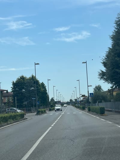 The image shows a straight, paved road in Castiglione delle Stiviere, Italy, with a clear sky and scattered clouds. The road is lined with trees, streetlights, and some buildings on either side. Several cars are visible on the road, some moving and some parked. There are also pedestrian crossings marked on the road. The overall scene depicts a typical suburban or semi-urban environment in Italy.