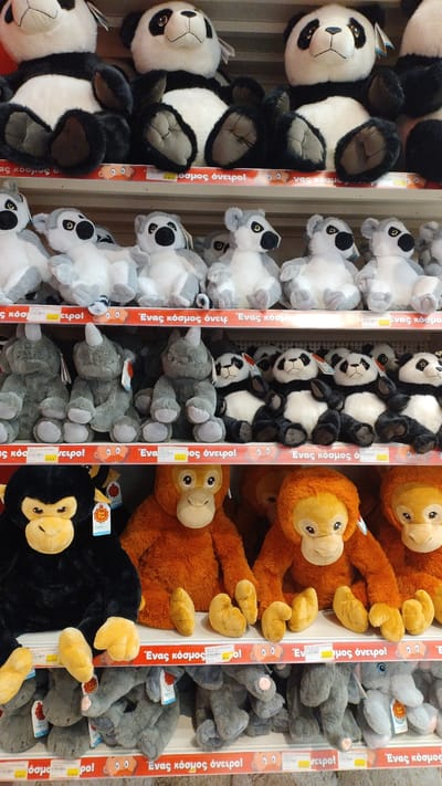 The image shows a retail display of stuffed animals in a store. The stuffed animals are arranged on shelves and include pandas, monkeys, orangutans, and elephants. The store appears to be a toy store or a department store with a toy section. The lighting is bright and evenly distributed, suggesting an indoor setting. The shelves are made of metal and are filled with plush toys. There are price tags visible on some of the toys, which are in Euros, suggesting the location is within the Eurozone. The writing on the price tags and promotional materials appears to be in Greek. The specific location is the Jumbo store in Pylaia Municipal Unit, Greece, as indicated by the Greek text which translates to "Jumbo is a world of dreams".