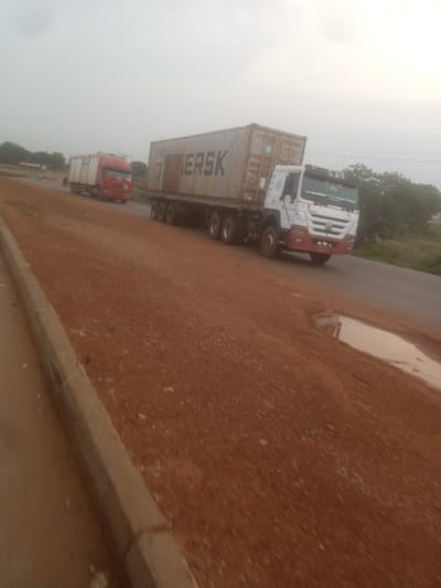A wide, unpaved road with a distinct reddish-brown dirt surface is visible, bordered by a concrete curb on the left. In the background, two large trucks are traveling on the paved portion of the road. The truck on the right is a white semi-truck with a large, beige shipping container that has the word "MAERSK" printed on its side in black capital letters. The truck has a red and white cabin. The truck on the left is a red semi-truck with a white trailer. The sky is overcast, creating a hazy, diffused light. Trees and some low vegetation are visible on the sides of the road. There is a large puddle of murky water on the paved road near the white truck. The overall scene suggests a dusty, rural environment, possibly in Nigeria.