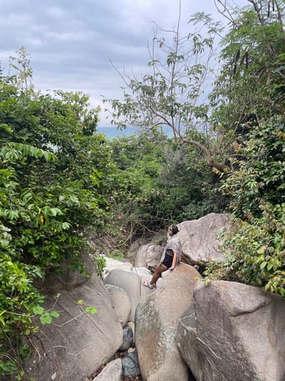 The image depicts a natural, densely vegetated environment in Ninh Hòa, Vietnam, featuring a streambed composed of large, smooth grey boulders under an overcast sky. A single female individual is seated on one of these boulders in the midground, angled slightly away from the camera, looking upwards and to her left. She wears a light-toned, possibly plaid, shawl or wrap over a dark top, dark shorts, and light pink sandals. A dark mobile device is held in her right hand. White, turbulent water flows between the boulders at the base of the scene. Lush green trees and bushes frame the area, with a narrow view of a distant blue sea on the horizon, visible above the vegetation.