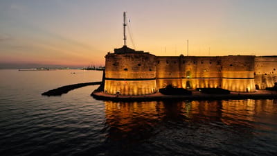 This media file depicts an aerial view of the Aragonese Castle in Taranto, Italy, at sunset. The imposing stone structure, illuminated by warm golden lights, stands proudly on a small island surrounded by calm, dark water. The castle's ancient walls, punctuated by small windows and battlements, create a dramatic silhouette against the dusky sky. A tall antenna mast rises from the highest point of the castle, reaching towards the colorful twilight.
The sky transitions from a pale blue at the top to a vibrant orange and yellow near the horizon, casting a warm glow over the scene. Faint reflections of the castle's lights shimmer on the water's surface, adding to the serene and romantic atmosphere. In the distance, the outlines of a port with cranes and ships are visible, hinting at the city's maritime significance. The overall mood is one of historical grandeur and tranquil beauty, capturing a moment of peaceful transition between day and night. No people are clearly visible. The text "Taranto, Italy" is not visible in the image.