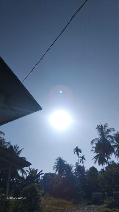The image depicts a bright, sunny day with a clear blue sky. The sun is prominently featured, emitting strong rays and lens flare. In the foreground, a section of a corrugated metal roof extends from the left, casting a shadow. Below and to the right of the roof, lush green foliage and various trees, including palm trees, are visible. A thin, dark wire stretches diagonally across the upper portion of the sky. The bottom left corner of the image displays the text "Galaxy A05s". The scene appears to be in a tropical or subtropical environment, consistent with a location in Rambongpayong, Indonesia. There are no people or animals visible in the image. The overall impression is of a natural outdoor setting bathed in intense sunlight.