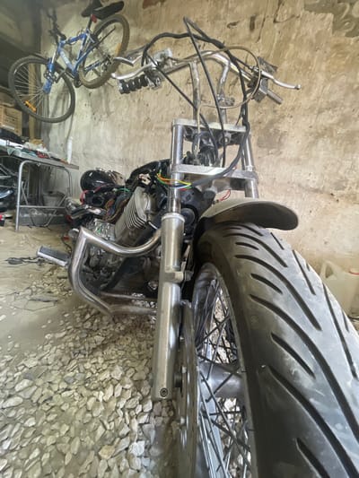 In a garage in Znamensk, Russia, a partially disassembled motorcycle is the main subject. The motorcycle is positioned at a low angle, with its front wheel and engine prominent. Exposed wiring is visible around the engine, suggesting ongoing maintenance or customization. To the upper left, a blue bicycle hangs from the ceiling. Various tools and what appear to be storage boxes are present on a metal cart in the background. The floor is covered in gravel. The walls of the garage are made of rough, textured concrete.