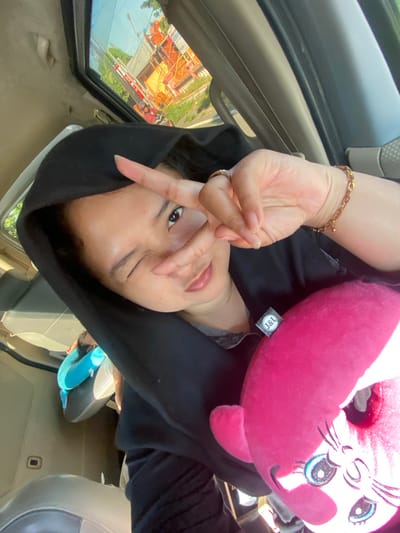 A young Asian woman is pictured from the chest up, taking a selfie inside a vehicle. She is wearing a black hooded garment, and her left eye is winking. Her right hand is raised, forming a peace sign over her face. She is holding a large, bright pink plush toy with cartoonish blue eyes and whiskers. A gold-colored bracelet is visible on her right wrist, and a ring is on her left index finger. The interior of the vehicle, including beige-colored seats and a sunroof, is visible. Through the sunroof, an outdoor scene shows a building with orange scaffolding and a red and white barrier, suggesting a construction site or road closure. The presence of a motorcycle with a rider outside the vehicle indicates a street environment. The scene is likely located in Gayaman, Indonesia, given the context of Indonesian postal services potentially related to the "J&T" label on the plush toy.