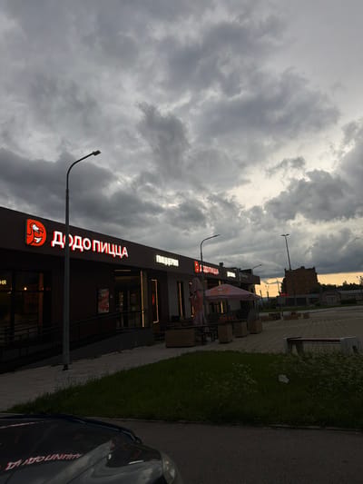 The image shows a cloudy sky over a commercial building in Chusovoy, Russia. The building has a prominent sign in red neon letters that reads "ДОДО ПИЦЦА", which translates to "Dodo Pizza". To the right of this sign, another sign in white letters reads "ПИЦЦЕРИЯ", meaning "Pizzeria". There is a street light in front of the building. The area in front of the building features a paved walkway and a grassy area with a bench. A parked car is partially visible in the foreground, with its reflection showing in the car's hood. The overall scene suggests an overcast day in an urban or suburban setting.