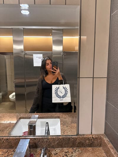 The image depicts a female individual taking a mirror selfie in a public restroom. The location is a restroom within a commercial or public facility in Lawrence Township, United States; specific address not discernible.
**Individual:** A female with dark, shoulder-length hair and sunglasses atop her head is visible from the chest up, reflected in the mirror. She is wearing a black V-neck top, a dark brown or black open cardigan, and dark lower garments. She holds a dark-colored smartphone with a patterned case in her right hand, positioned to capture her reflection. In her left hand, she holds a small white shopping bag with dark blue or black print featuring a "C" enclosed by a laurel wreath and the word "PINK" below.
**Environment:** The restroom features a speckled light brown and black granite or stone countertop with a white rectangular sink basin. A modern stainless steel faucet and a second fixture (possibly a soap dispenser) are installed on the countertop. Reflected in the mirror are two stainless steel toilet stalls with light brown paneling above. A white toilet bowl is partially visible within the leftmost stall. A white sign with inverted black text, possibly indicating "Temporarily Closed," is affixed to the middle stall door. Recessed ceiling lights and additional warm-toned linear lighting fixtures are visible in the reflection. The wall to the right of the mirror features large beige rectangular tiles and a section of darker, textured vertical panels.