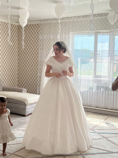 The image depicts a bride in a white, sparkling wedding dress standing indoors, possibly in a residential setting. She is wearing a veil and a tiara. Beside her, a young child in a white dress stands barefoot on a patterned rug. The room is decorated with white balloons and ribbons hanging from the ceiling. A sofa and a window with lace curtains are visible in the background. The lighting suggests daytime. The location is identified as Astrakhanka, Kazakhstan.