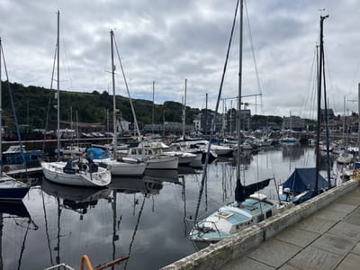 The image displays a marina or harbor in Douglas, Isle of Man, under an overcast sky. Numerous sailboats and some motorboats are moored in calm, dark water, which provides clear, mirrored reflections of their hulls, masts, and rigging.

In the mid-ground, a white sailboat with blue trim features the name "PRIDE OF MANN" prominently displayed in blue lettering on its stern cover. In the foreground on the right, a light blue and white sailboat with "CORSA" on its bow and the number "5017" on its hull is visible, partially obscured by a concrete quay. This boat has a blue canvas cover over its cockpit. The quay itself is made of light grey concrete paving stones and a low concrete wall.

The background on the left is dominated by a lush, green hillside covered with trees. Below the hillside, industrial and commercial buildings are visible, including one with a partially discernible red "WEDGWOOD" sign. A parking lot with multiple vehicles is adjacent to these structures. Further right in the background, a mix of multi-story modern and traditional buildings line the waterfront, with a yellow construction crane visible above them. No individuals are clearly discernible in the scene.