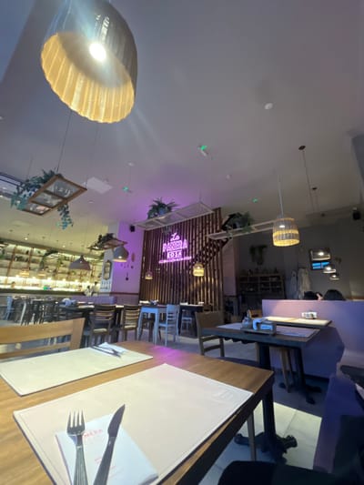 This indoor shot depicts a restaurant interior with a focus on dining tables set with placemats, cutlery, and napkins. The ambiance is illuminated by several pendant lights, including a large, woven one dominating the lower left of the frame. Overhead, there's a complex arrangement of decorative elements including hanging plants and framed artwork.
The restaurant, "La Panera Rosa Cocina Casera Non-Stop," is identifiable by signage in the background. The location is likely Lomas de Zamora, Argentina, given the geotag. The scene is sparsely populated, with hints of other diners in the distance.