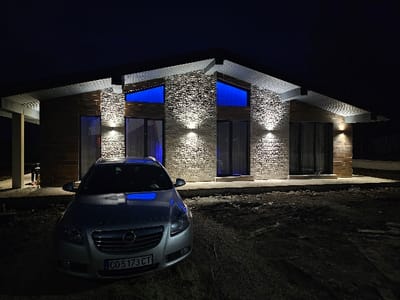 The image depicts a modern, single-story house with an angular roofline illuminated at night in Dragushinovo, Bulgaria. A silver Opel Insignia vehicle is parked on an unpaved, dark dirt foreground, facing the house. The house's facade features sections of light-colored textured stone or brickwork centrally, flanked by darker horizontal paneling. Multiple wall-mounted lights cast warm upward and downward illumination onto the exterior walls. Two rectangular windows on the upper central facade emit a distinct blue light from within, with this blue glow reflecting on the car's hood. The vehicle's front license plate is visible, displaying 'CO 5173 CT'. The sky is completely dark, indicating nighttime. No individuals, actions, or interactions are present in the scene.