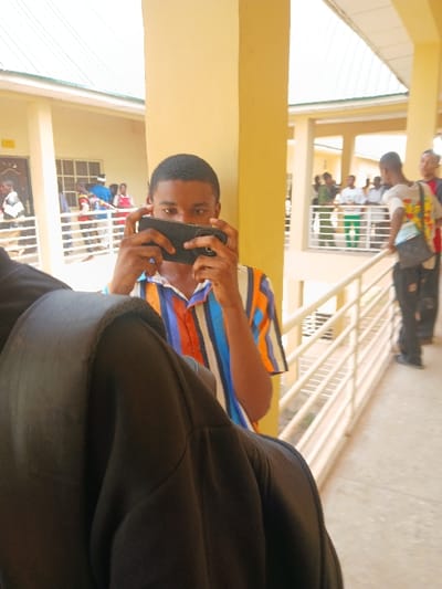The image depicts a daytime scene within a multi-story building complex in Ugi Majowel, Nigeria. The architecture features light-yellow painted walls and pillars, white metal railings, and a light-colored sloped roof.
In the foreground, the left shoulder and upper back of an unidentified individual, wearing a dark-colored garment, partially obscure the lower left quadrant of the frame. Centrally positioned behind this obstruction, a young male subject, likely in his late teens or early twenties, holds a black smartphone horizontally at eye level with both hands, appearing to record or photograph. He has short dark hair and wears a horizontally striped short-sleeved shirt with blue, white, and orange segments. His eyes are focused forward, visible above the phone.
To the subject's right, a prominent light-yellow structural pillar stands. Beyond this pillar and to the right, a walkway with white metal railings extends into the background. Several other individuals are visible on this walkway; one young male with a backpack is leaning on the railing, looking towards the left. Additional figures are observed further along the walkway and in a background structure to the left, which has windows and doorways, also featuring railings and visible occupants. The lighting suggests an outdoor or semi-outdoor environment under natural light.