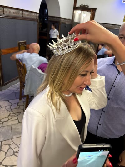 In Zavod Mikhailovskii, Russia, a woman with blonde hair is having a tiara placed on her head by a person whose hand is visible in the frame. The woman is wearing a white blazer over a black top and a pearl necklace. She has red nail polish. A man in a blue shirt is partially visible on the right side of the frame, and another man with a bald head is seated in the background on the left. The environment appears to be an indoor venue with tiled flooring and decorative wall panels.