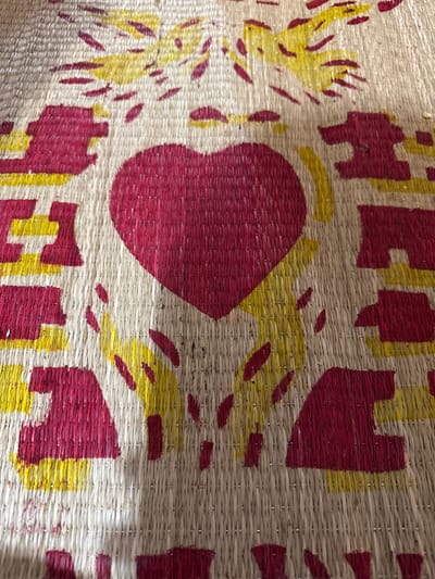 A close-up, overhead view displays a light-beige woven mat with a patterned design in red and yellow, observed in Ko H'Neh, Vietnam. Centrally, a large red heart is depicted. Below and to the sides of the heart are stylized yellow shapes resembling cupped hands or supporting elements, with additional yellow and red abstract shapes above, suggestive of wings or radiating energy. Blocky red letters with yellow outlines are positioned around the central heart, forming the word "LOVE"; specifically, the letter "E" is visible to the left, and the letter "V" is visible to the right. The mat exhibits a distinct horizontal and vertical weave, showing minor textural variations. No individuals, actions, or further environmental context beyond the mat itself are visible within the frame.