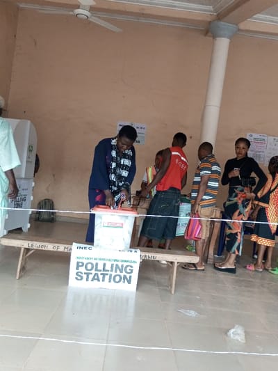 The image displays an indoor polling station in Ajalli, Nigeria. At the center, a person dressed in a dark blue garment and patterned scarf is positioned over a white plastic ballot box with a red lid, appearing to cast a vote or interact with the ballot system. The ballot box rests on a wooden bench, and its label features the Nigerian flag. To the left, a white polling booth marked "POLLING ZONE" is visible, with a person in light attire standing beside it. To the right, a line of four individuals stands waiting: a male in a red sleeveless shirt, a male in a striped polo shirt carrying a striped bag, a female in a black top and patterned skirt holding a black handbag, and a partially visible female in a patterned dress.
A white sign on the tiled floor in the foreground reads "INDEPENDENT NATIONAL ELECTORAL COMMISSION POLLING STATION" and "INEC EC 30 A." A white string marks a line on the floor. The walls are light brown/beige, exhibiting minor cracks, and have several official documents affixed, one marked "EC 30 C." A white ceiling fan is mounted above, and a concrete pillar supports the ceiling on the right. A green backpack is on the floor to the left of the bench. Wires and electrical equipment are visible on a surface behind the ballot box.