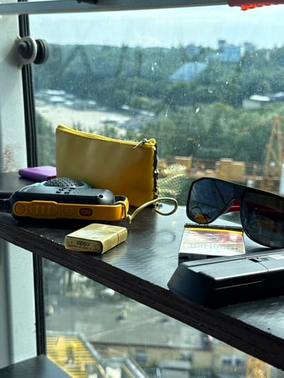 The image shows a collection of personal items placed on a dark wooden shelf in front of a window with a view of an urban landscape. The items include a yellow pouch, a black and orange walkie-talkie, a brass Zippo lighter, a pack of Marlboro cigarettes, a pair of black sunglasses, and a black electronic device, possibly a remote control. The background reveals a blurred cityscape with trees and buildings, suggesting an outdoor or elevated vantage point. The lighting suggests it is daytime. The image does not provide any specific clues to pinpoint the location within Gdynia, Poland, other than a general urban setting. There are no people or animals depicted in the foreground or midground.