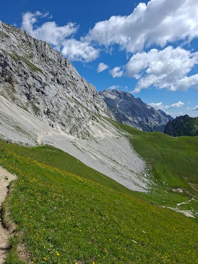 The image captures a vast alpine landscape in Mieming, Austria. In the foreground, a dirt path winds through a lush green meadow dotted with numerous yellow wildflowers. The meadow slopes downwards and to the right, eventually transitioning into a steep, rocky mountainside. This mountainside dominates the left side of the frame, its grey, rugged surface interspersed with patches of green vegetation. A narrow, winding trail is visible snaking its way up this rocky incline, and several small figures, likely hikers, are discernible on this path. Further in the background, a series of majestic, jagged mountain ranges recede into the distance under a bright blue sky populated by fluffy white clouds. The overall scene conveys a sense of natural grandeur and an invitation to explore the mountainous terrain.