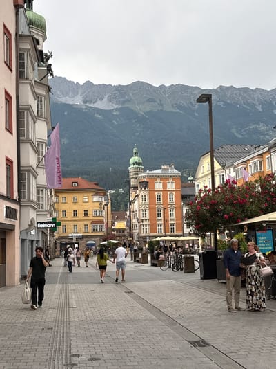 The image depicts a street scene in Innsbruck, Austria, with a prominent mountain range in the background. The photograph is oriented vertically. The street is paved with cobblestones and lined with multi-story buildings featuring various architectural styles, some with colorful facades and decorative windows. A few people are visible walking along the street, and some are seated at outdoor cafes.

One man in a dark t-shirt and pants is walking with a bag over his shoulder, talking on his phone. Further up the street, a couple stands near a flower-laden trellis, examining what appears to be a menu or brochure. A man in a blue shirt and khaki pants stands nearby, looking towards them.

Signs are visible on some buildings, including one with blue lettering that reads "Stadt der Liebe mag ich!" and another with green and white lettering that says "Aelmann". A purple banner with white text, which appears to read "FRAUENLAUF", is draped across a building. The time of day appears to be daytime, possibly late afternoon, given the lighting. The weather seems overcast. The presence of the mountains and the architecture strongly suggest the location as Innsbruck.