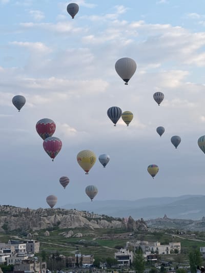 The image shows a multitude of hot air balloons floating in the sky above Göreme, Turkey. The balloons are of various colors and patterns, and some appear to be clustered together. In the background, there are buildings and rock formations typical of the Cappadocia region. The sky is overcast with a mix of clouds, suggesting a slightly hazy atmosphere.