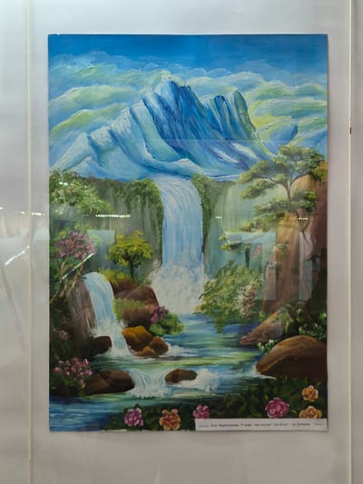 The image shows a painting of a landscape featuring a waterfall cascading down a rocky mountainside. The left side of the painting depicts a large, imposing mountain with blue and white hues, suggesting snow or ice. The waterfall is the central element, flowing from the higher elevations through greenery and trees. Rocks and vegetation are interspersed along the waterfall's path. Flowers bloom on the right side of the painting. The painting is framed and has a paper label attached to the top right corner with Bulgarian Cyrillic text: "Ема Бориславова-9 клас. Ару ателие" Дон Кеш" гр. Дупница". The painting is likely displayed behind glass or plastic, as there are reflections visible on its surface. The location in the city of Srebarna, Bulgaria, is not directly depicted in the image and cannot be inferred from the available visual information.