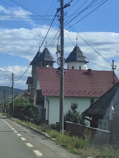 The image depicts a street scene in Rona de Jos, Romania, dominated by a church with distinctive twin spires. The church features a prominent red tiled roof, and one of its spires is topped with a cross. In the foreground, a paved road runs along the left side of the frame, marked with dashed white lines. Electrical poles and a dense network of overhead wires are a significant feature of the scene, crisscrossing the sky above the church and the road. Adjacent to the road, there is a grassy verge with a low stone curb and a wooden fence. Behind the fence, vegetation and parts of houses are visible. The background shows rolling hills under a blue sky with scattered white clouds.