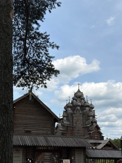 In the city of Nevskii Parkleskhoz, Russia, a vast pine tree trunk occupies the left foreground of the image, partially obscuring a wooden building with a gabled roof and intricate carvings. Behind this structure, a magnificent wooden church with numerous domes and crosses stands prominently against a bright blue sky dotted with white clouds. The church exhibits traditional Russian architecture, characterized by its multiple onion domes of varying sizes, each topped with a cross. The overall scene depicts a serene outdoor environment, likely a historical or cultural site, with the natural elements of the tree and sky complementing the man-made wooden structures.