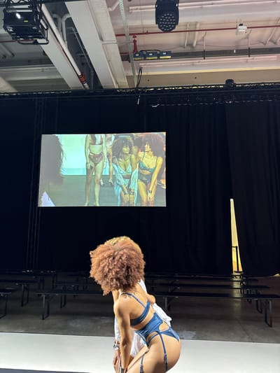 The image depicts an indoor scene at what appears to be a fashion event in Issy-les-Moulineaux, France. In the foreground, a person with voluminous reddish-brown curly hair is viewed from behind, bending forward and to the left. This individual is wearing a teal blue one-piece lingerie garment featuring garter straps and a bra-like top. A white garment or accessory is partially visible on their left arm.

In the mid-ground, a large projection screen displays multiple female models in various lingerie and swimwear. From left to right on the screen, visible models include: one partially seen in dark swimwear, one in a red patterned two-piece, one with curly hair in a light blue sheer lace two-piece, and one with curly hair in a dark green two-piece. Behind the foreground subject and in front of the screen, rows of empty, dark-colored benches are arranged, suggesting an audience seating area.

The background consists of black theatrical drapes spanning the width of the space behind the screen and along the right side, with a narrow vertical opening on the right revealing a light source. The ceiling reveals industrial infrastructure, including white pipes, metal cable trays, and multiple stage lighting fixtures, one on the far left and another prominent black fixture with multiple LED lights on the right.