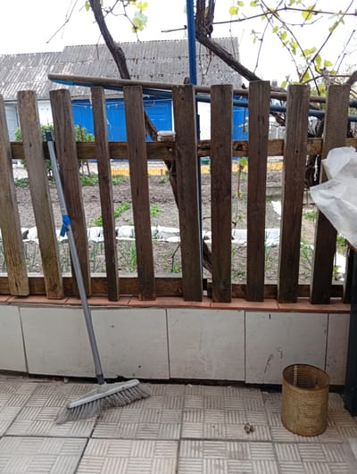 The image depicts a scene likely on a porch or patio. In the foreground, a broom leans against a weathered wooden fence. A cylindrical tin can rests on the tiled floor. Beyond the fence, cultivated land is visible, possibly a garden. A house with a grey, corrugated roof and a blue door is in the background. The location is identified as Kostopil, Ukraine. There are no people visible in the image.