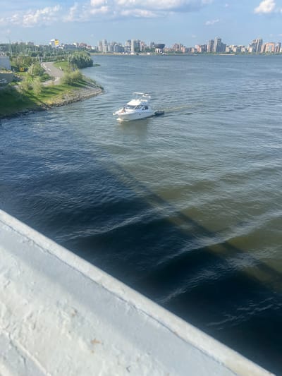 The image captures a wide shot of a body of water, likely a river or bay, with a city skyline in the background. In the foreground, a white railing or bridge structure is visible. The water is choppy with visible waves, and a shadow is cast across it, suggesting the viewpoint is from above. A white motorboat with a small inflatable dinghy in tow is moving across the water. On the left side of the frame, the shore is lined with green vegetation and a paved path, possibly for pedestrians or cyclists. Further inland, trees and various buildings, including tall structures and lower-rise buildings, form the cityscape. The sky is partly cloudy with a clear blue sky visible. The scene appears to be a sunny day. The location is identified as Дачное, Russia.
