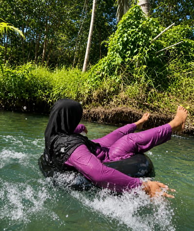 A person wearing a black hijab and purple attire is tubing down a river in Silaiyajae, Indonesia. The person is seated in a black inner tube, with their legs extended and bare feet visible above the water. Water is splashing around the inner tube, indicating movement. Lush green vegetation and trees line the riverbanks, with a large, ivy-covered tree dominating the right side of the frame. The water is clear and green, reflecting the surrounding foliage. Sunlight illuminates the scene, casting shadows and highlighting the water's movement.