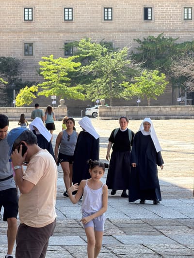 The image depicts a scene in the courtyard of El Escorial, located in San Lorenzo de El Escorial, Spain. In the foreground, a man wearing a blue cap and a light-colored t-shirt is on the phone. To his right, a young girl in a white tank top and light purple shorts walks past. Behind them, several women in nun habits are walking. One nun, in a black habit and white veil, is facing forward. Another nun, also in a black habit and white veil, is walking behind and to the left of the first. To the left of these nuns, another woman in a dark habit and white veil is visible. Further back, two women in casual clothing are walking away from the camera, and two other figures are partially visible. The background features a large stone building with multiple windows, and green trees. The ground is paved with large, grey stone slabs.