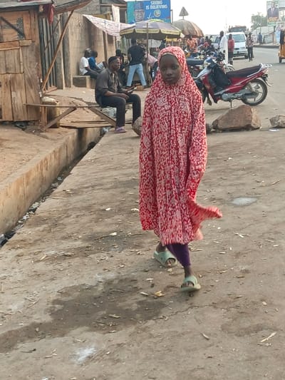 An outdoor scene in Alizaga, Nigeria, depicts a street with pedestrian and vehicle activity. In the immediate foreground, a young person, possibly female, stands or walks toward the viewer. They wear a red and white patterned full-length garment covering the head and body, purple trousers visible beneath the dress, and light green slide sandals. Their gaze is directed forward.
To the left, a concrete sidewalk or raised platform features a shallow open drainage ditch filled with debris. Adjacent to this, crude wooden structures and a concrete wall are visible. A man in a dark shirt, dark trousers, and a dark cap is seated on a makeshift wooden bench alongside the ditch, holding a mobile phone. Further back on the left, other individuals are seated or standing near structures.
The right side of the image shows a paved road. A red motorcycle is parked on the shoulder, with large rocks positioned nearby. In the mid-ground, a triangular street sign is visible. Further down the road, additional vehicles, including cars and trucks, are present, alongside various pedestrians and street vendors operating under umbrellas. A prominent billboard in the background displays text including "SEEING IS BELIEVING" and the image of a political figure. The ground throughout the visible area is primarily concrete, appearing dusty and worn, with scattered debris.