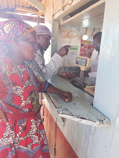 This image captures a lively scene at a butcher stall in Kaplelwo, Kenya, under bright daylight. Three individuals are present, engaged in a transaction.
In the foreground, a woman is seen from the chest up, wearing a vibrant red, blue, and white patterned dress and a matching headscarf. She has a warm smile on her face and is looking towards the butcher. Her left arm rests on the weathered wooden counter that separates the customers from the stall.
Beside her, slightly behind, a man wearing a light-colored hat and a grey long-sleeved top is visible in profile. He is extending his right arm and pointing towards something inside the stall, likely indicating a cut of meat or confirming a detail of the purchase.
Behind the counter, a butcher in a white coat is attending to the customers. Cuts of meat are visible on the counter, next to a weighing scale. The stall itself appears to be a simple, wooden structure, typical of a local market setting.
Notable details include a sign inside the stall displaying the "M-PESA" logo and a "TILL NUMBER: 269991," indicating the acceptance of mobile money payments. Below the counter, a poster featuring "MANCHESTER UNITED" and images of football players is visible. The worn, paint-peeled wooden counter adds to the authentic market aesthetic. The bright sunlight suggests it is daytime.