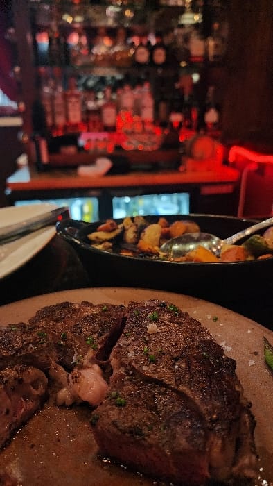 The image depicts a close-up, low-light scene within what appears to be a restaurant or steakhouse. In the foreground, a large, dark-brown steak, seasoned with coarse salt and finely chopped green herbs, is presented on a light-colored, possibly ceramic or wooden, plate. The steak has been cut into several pieces, revealing a medium-rare interior. Visible juices collect around the meat on the plate.
To the left and slightly behind the steak, a portion of a white plate with resting silverware (a fork and knife) is discernible. Centered in the midground, a black serving dish contains a medley of roasted vegetables, including what appear to be cubed potatoes, carrots, and possibly Brussels sprouts or other green vegetables. A silver serving spoon is partially submerged within the vegetables.
The background is significantly blurred, indicating a shallow depth of field. It reveals a dimly lit bar area with numerous liquor bottles arranged on shelves, illuminated predominantly by red ambient light. Below these shelves, a refrigeration unit with visible internal lighting is present. Red light strips are also visible along the bar counter. No individuals are visible in the scene. The specific location within the city of Jackson, United States of America, is not discernible from the visual information provided.