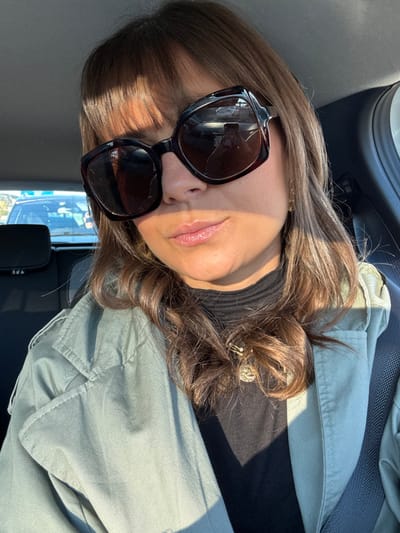 A woman poses in the sunlight, wearing large, dark sunglasses with a visible crack. She has brown hair styled with a decorative clip. She wears a black turtleneck under a light green jacket. The photo appears to be taken from an unusual angle, possibly from inside a car, with the side window visible in the background.
