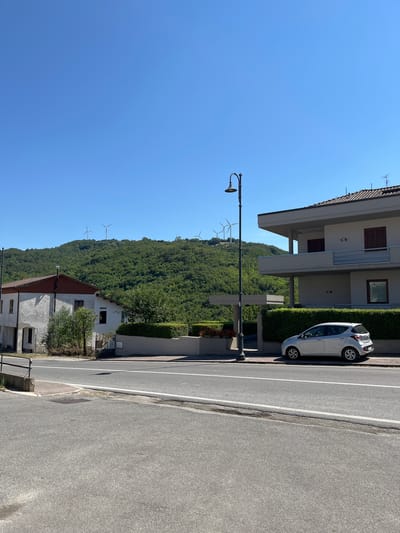 The image depicts a street scene in Foiano di Val Fortore, Italy, under a clear blue sky. On the left, an aged, white building with a reddish-brown roof is visible. Adjacent to it, a modern, multi-story building with a light-colored facade and dark-shuttered windows stands on the right. A silver car is parked on the sidewalk in front of the modern building. A street lamp with an ornate design is positioned between the two buildings, near the sidewalk. The background features a lush, green, tree-covered hillside with several wind turbines dotting the summit. The foreground shows a paved road with white lane markings.
