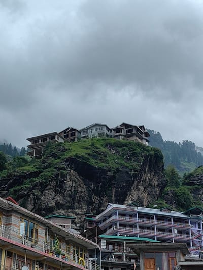 The image shows a view of Palchan, India, with buildings constructed on a steep, rocky mountainside under a cloudy sky. Several multi-story buildings are visible, some appearing to be hotels or guesthouses, with balconies and multiple windows. The architecture is modern, with some buildings appearing to be under construction or recently completed. The natural landscape features dense green vegetation covering the rocky terrain, with pine trees visible on the higher slopes. The sky is overcast, suggesting a potentially cool or damp climate. In the foreground, another building with a similar architectural style is visible, featuring balconies and rows of windows. The overall impression is of a developing tourist destination in a mountainous region.
