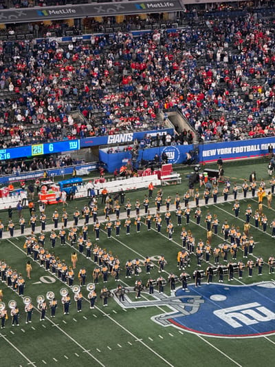The image presents an overhead view of MetLife Stadium in East Rutherford, New Jersey, United States, during a public event. A large marching band, comprising approximately 100 individuals, is arranged in organized formations across the green artificial turf playing field. Band members are uniformly attired in dark blue uniforms with gold accents and white shakos, carrying brass, woodwind, and percussion instruments. Several individuals in full gold-colored costumes are interspersed within the band formations.

The stadium stands, visible from the mid-level upwards, are densely populated with spectators, many wearing red and blue apparel. A scoreboard is partially visible in the upper left, displaying "12:16" and scores "25 6," with "GIANTS" and "NY" logos. "MetLife" branding is visible along the stadium's upper tier. The field features white yard lines, hash marks, and a prominent blue and gray New York Giants helmet logo painted on the turf. The sidelines contain various personnel, camera equipment on wheeled platforms, and a blue Gatorade cooler station.