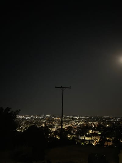 A nighttime cityscape of Xanthi, Greece, is depicted from a high vantage point, likely a hill overlooking the city. The sky is dark with a faint glow on the right, suggesting the presence of the moon, though it is obscured by haze. A wooden utility pole with crossbars stands prominently in the center of the frame, silhouetted against the night sky. Below the pole, the city sprawls out, a tapestry of illuminated buildings and streets. The lights create a warm, inviting glow that contrasts with the darkness of the sky. In the foreground, the dark silhouettes of trees frame the scene, adding depth and a sense of natural boundary to the urban landscape. The overall impression is one of a peaceful, slumbering city under the vast expanse of the night sky.
