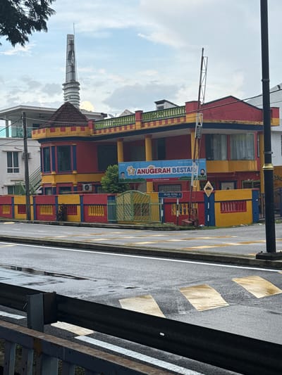 The image captures a scene in Ampang Jaya Municipal Council, Malaysia. A brightly colored building, predominantly red, yellow, and blue, stands adjacent to a road. A sign reads "ANUGERAH BESTA" in blue lettering. There's also a street sign indicating "JALAN PUTRA 4" and another sign indicating that the MADKA school is in the vicinity. In the background is a tall, white minaret and there is a small tree to the left of the minaret. The road appears wet, marked with yellow lines and a series of diamond-shaped markings. Part of a grey metal structure, potentially a bridge or railing, is visible in the foreground.